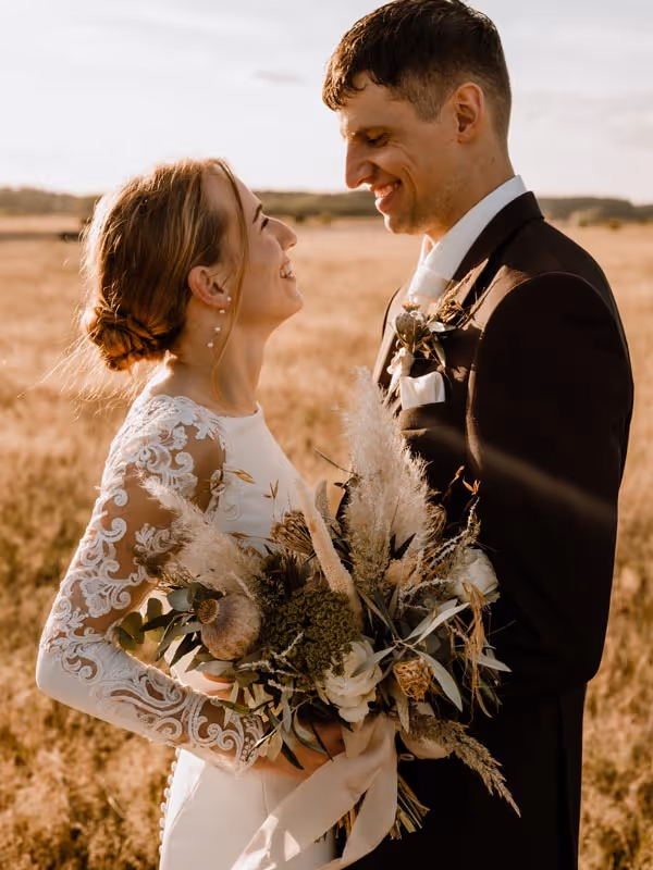 bruidspaar in graanveld lachend naar elkaar met tussen hen in het bruidsboeket