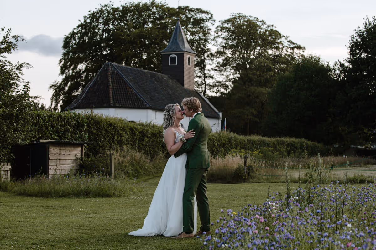 Fotoshoot bruidspaar, met een mooi wit kerkje op de achtergrond en een paar bloemenveld op de voorgrond